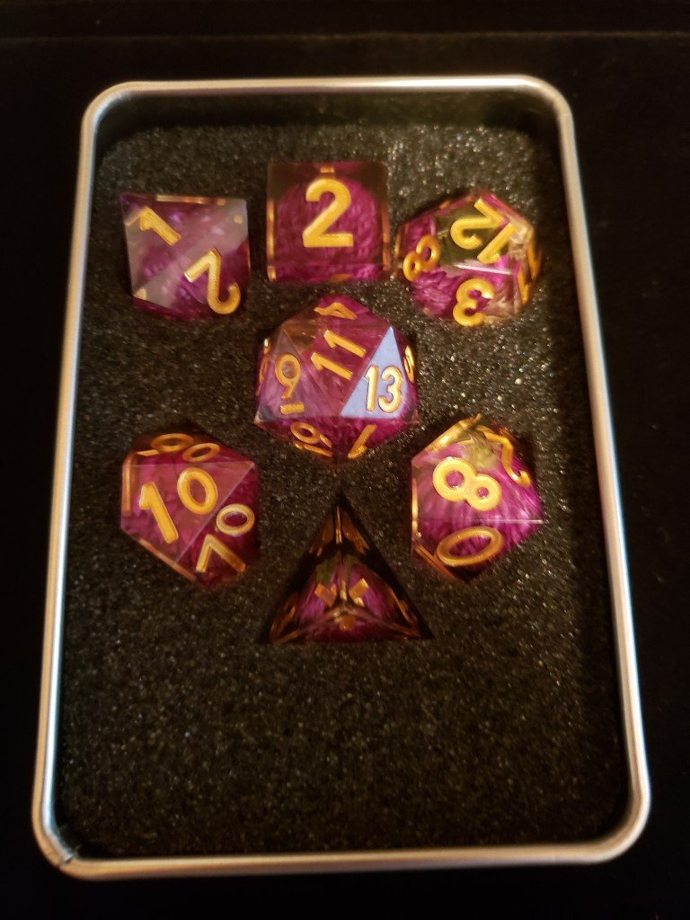A full set of Metallic Dice Game's Handcrafted Thousand Day Red. Clear dice with gold numbering and a violet flower in the center