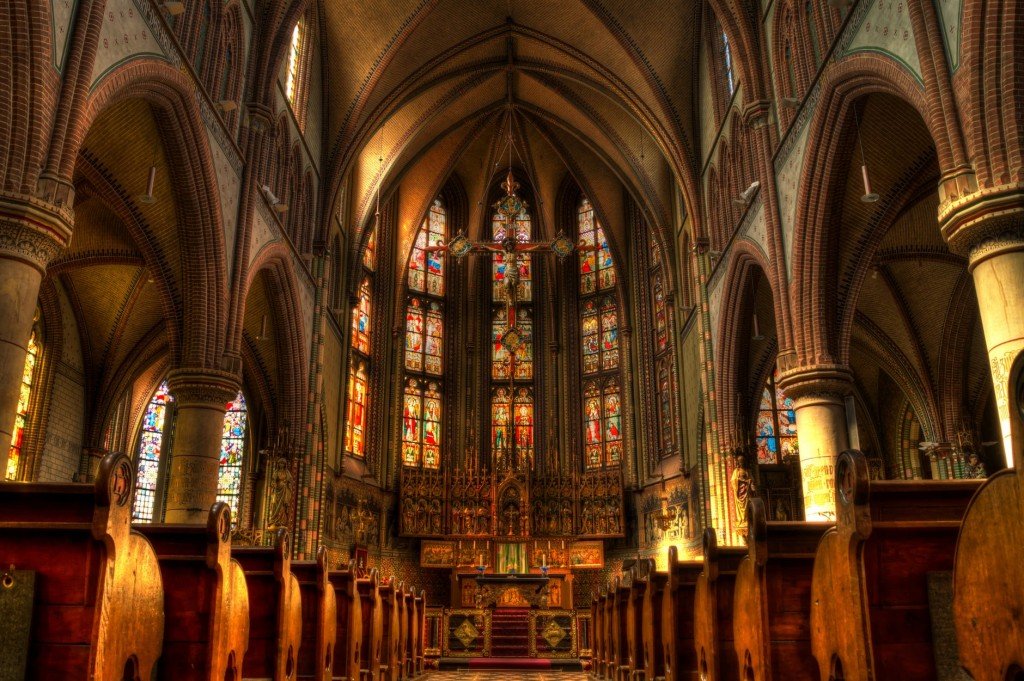 the inside of a catholic church. Rows of pews are on each side of the photograph and the altar is at the center of it surrounded by stain-glass windows